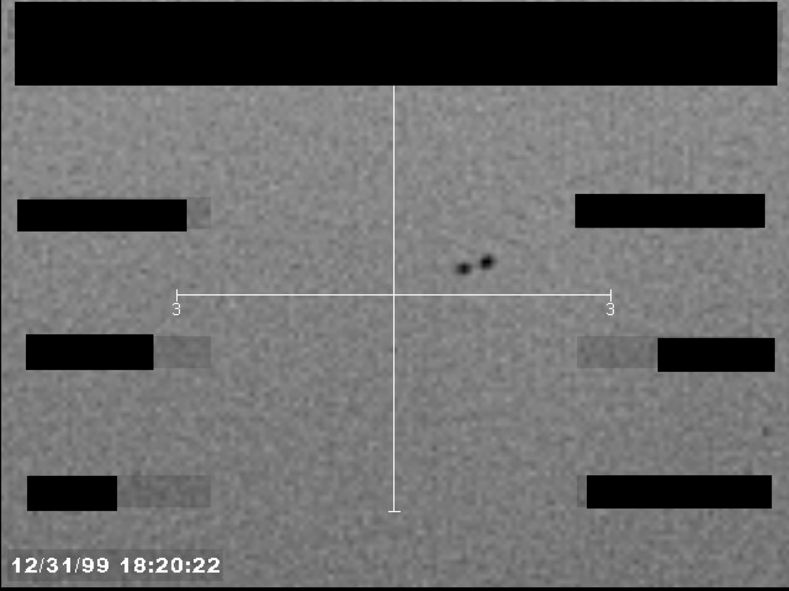 A grainy, black-and-white image from a sensor or camera, showing a crosshair centered on two small, dark objects. A timestamp is visible in the lower-left corner.
