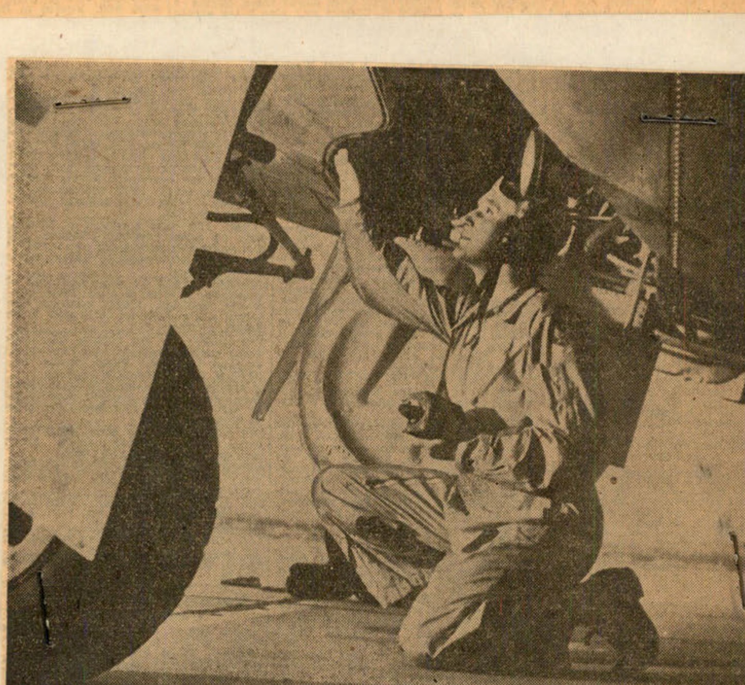 A man in a military uniform kneels under the wing of an airplane.