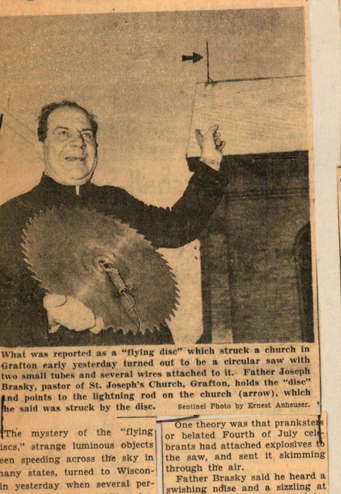 What was reported as a "flying disc" which struck a church in Grafton early yesterday turned out to be a circular saw with two small tubes and several wires attached to it. Father Joseph Brasky, pastor of St. Joseph's Church, Grafton, holds the "disc" and points to the lightning rod on the church (arrow), which he said was struck by the disc. -Sentinel Photo by Ernest Anheuser.