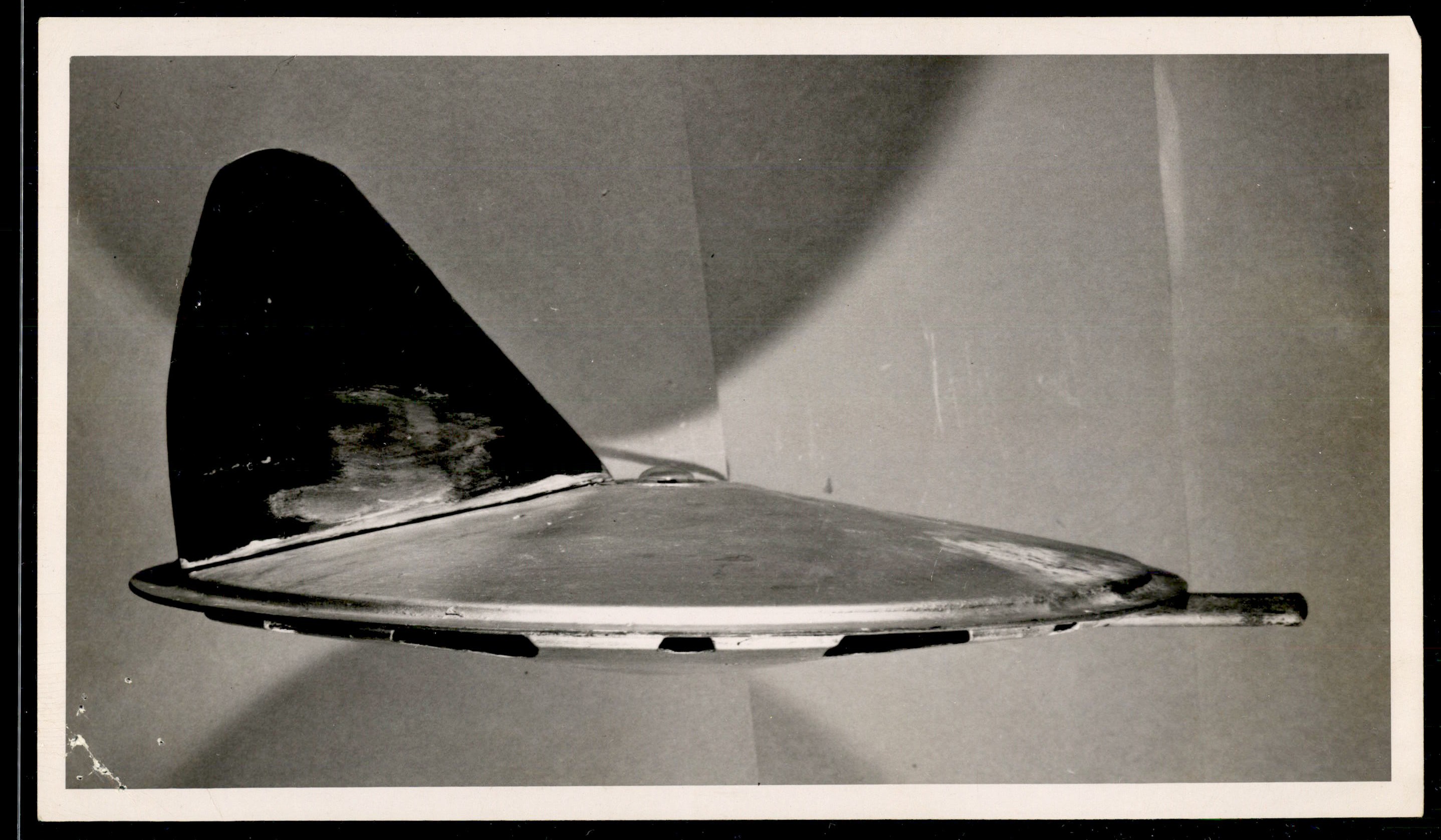 Side view of a metallic, disc-shaped object with a large vertical fin attached to the top.