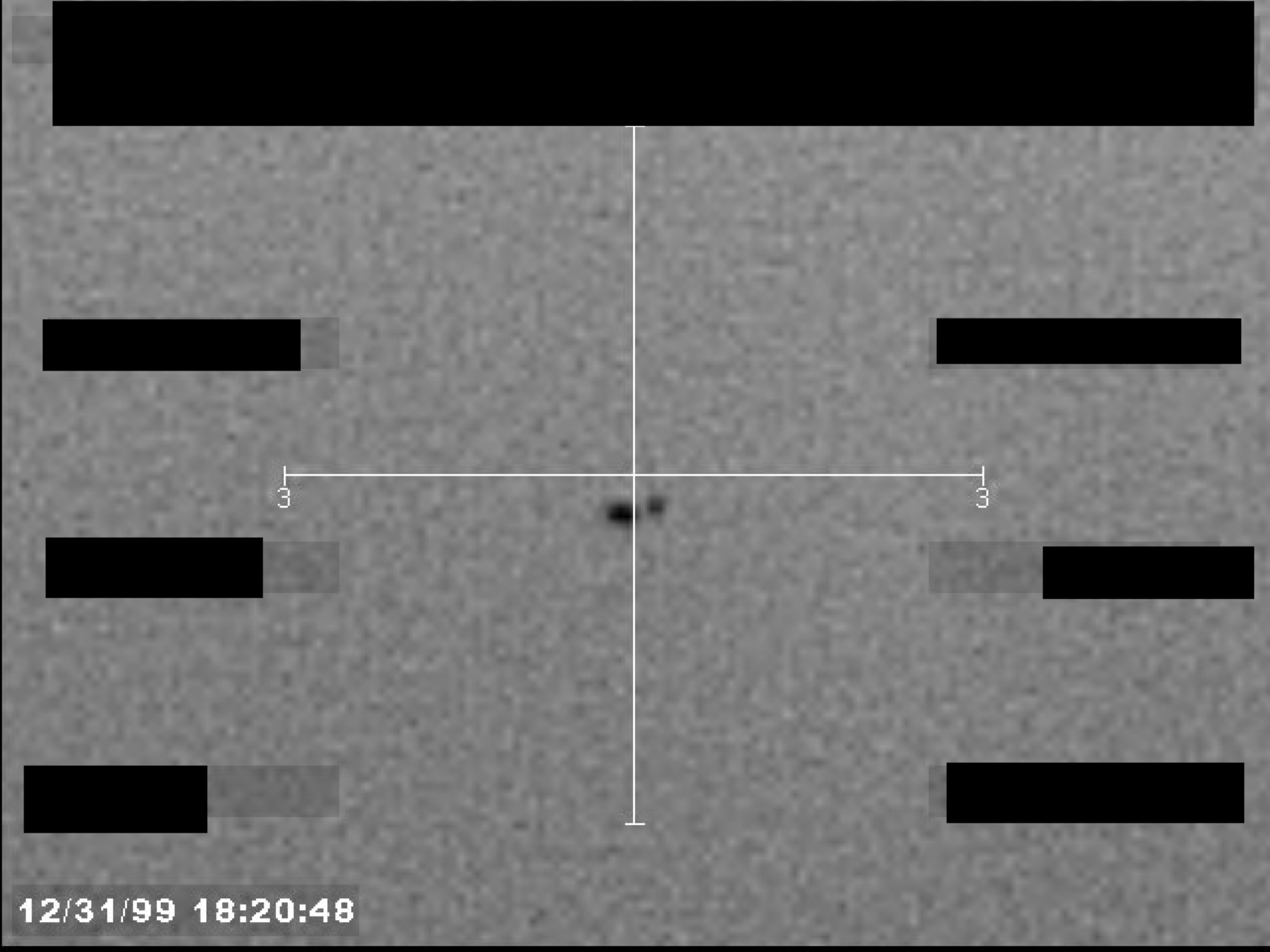 A grainy, black-and-white image, possibly from a sensor, showing a central dark object within crosshairs. A timestamp is visible in the lower-left corner, and several data fields are redacted.