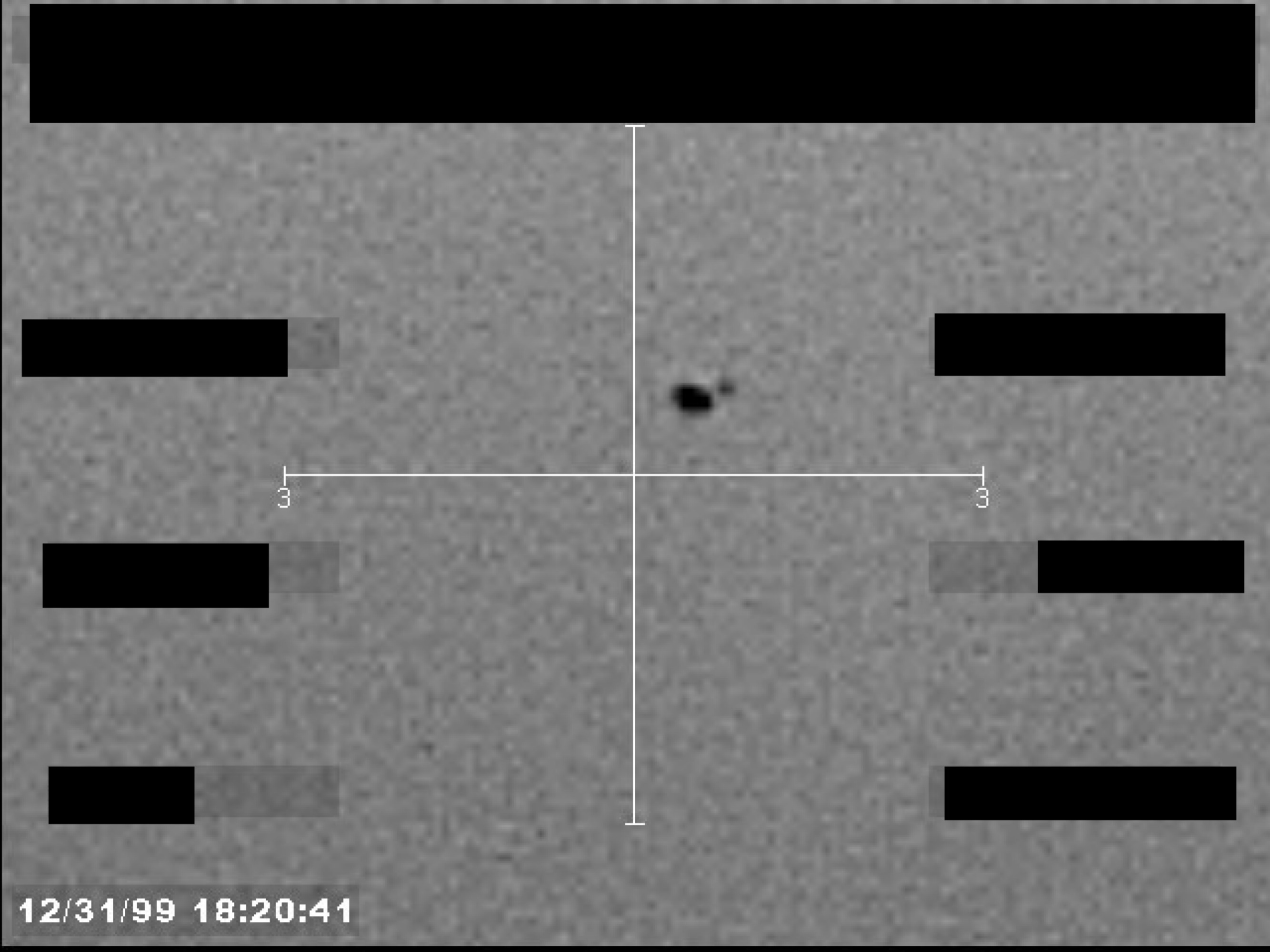 Sensor image, possibly infrared, showing a small, dark, irregularly shaped object at the center of a crosshair display. A timestamp is visible at the bottom left.